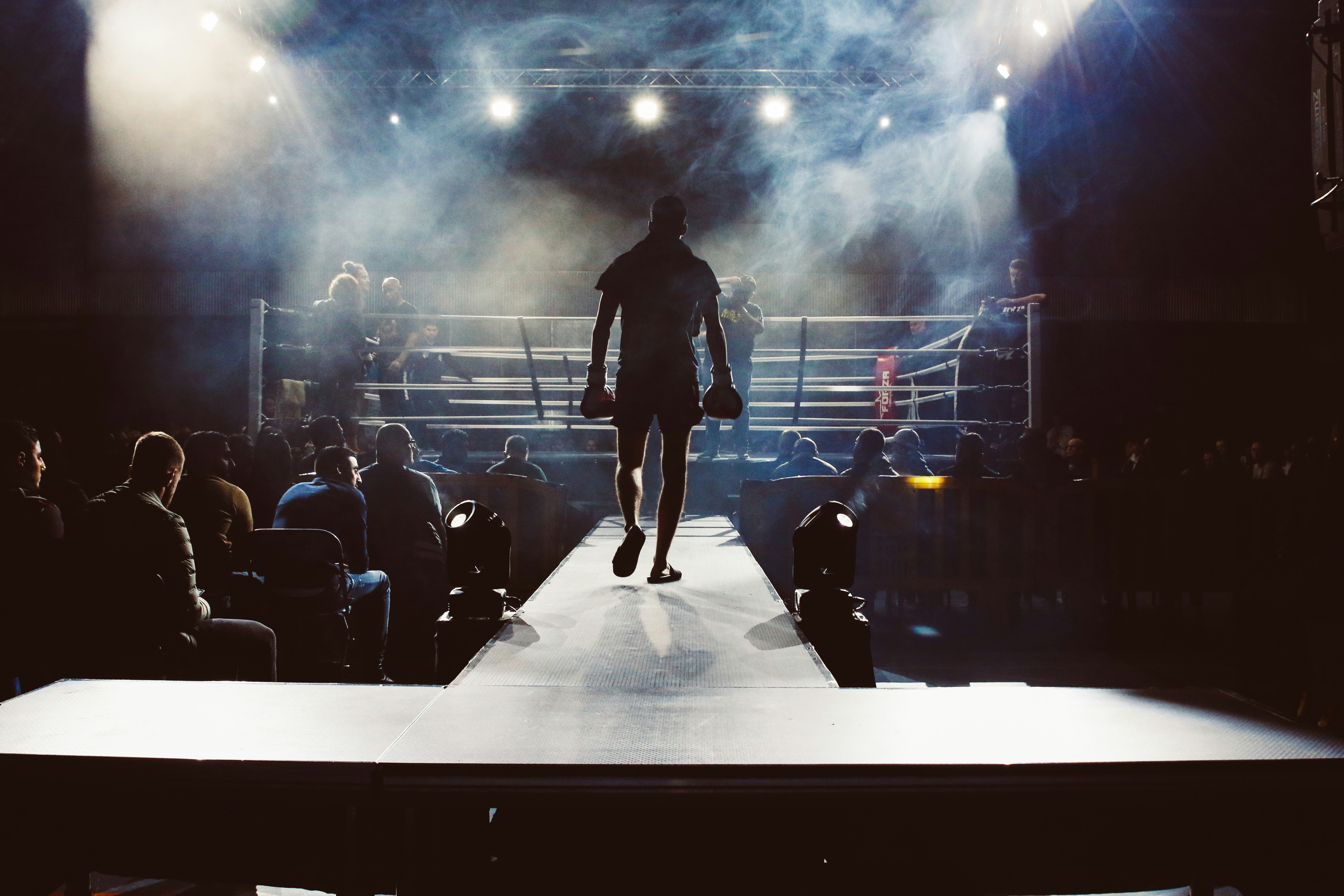 A lone boxer walking out to the ring through smoke and stage light, gloves in hand.