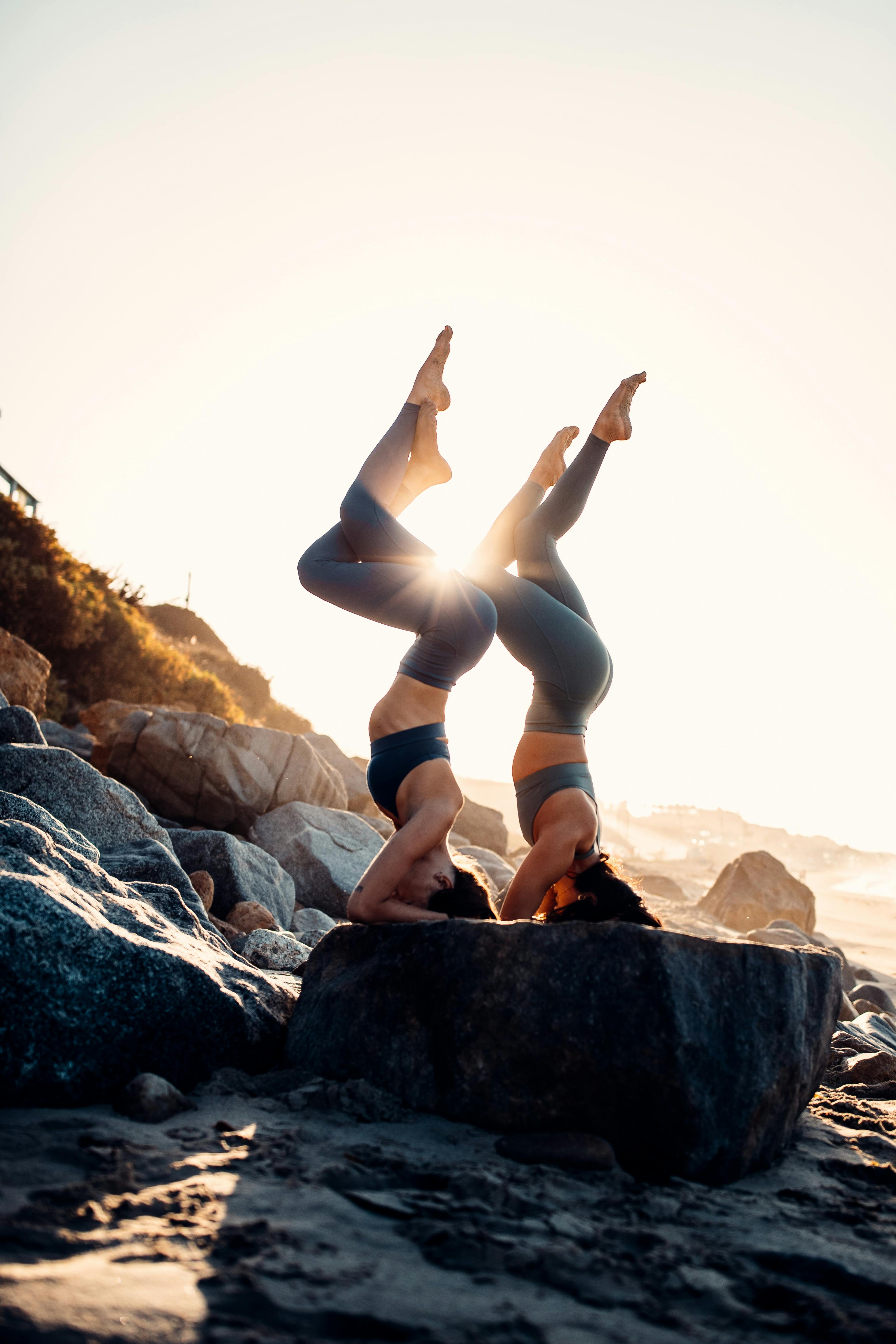 Two women in matching headstands at golden hour, on coastal rocks.
